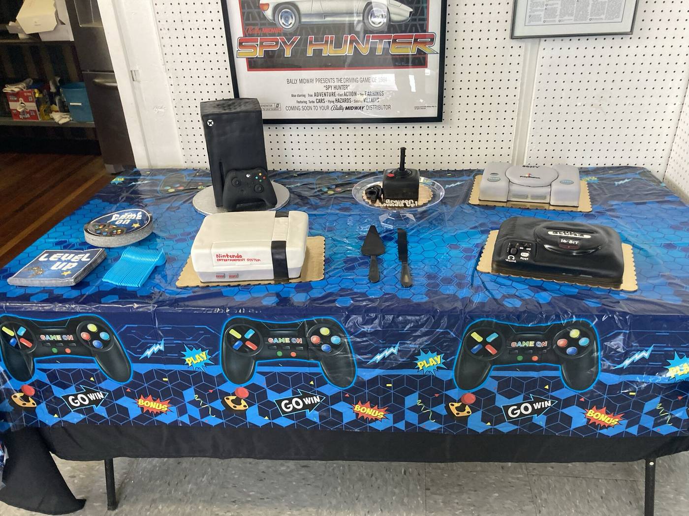 Birthday cakes shaped like an NES, Xbox, Sega Genesis and PlayStation on a video game tablecloth
