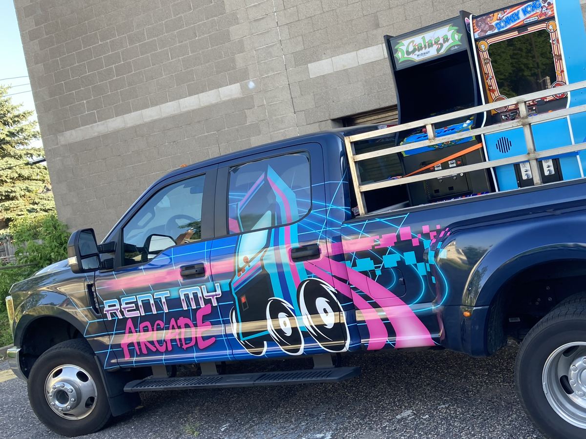 Rent My Arcade pickup truck wrapped in neon arcade graphics, hauling Galaga and Donkey Kong cabinets